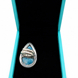 A silver ring with a teardrop-shaped blue stone inlay, displayed inside an open turquoise jewelry box with a black velvet lining on a white background.