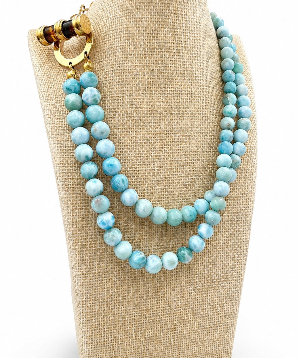 A double-strand beaded necklace with light blue marbled beads and a gold-toned clasp, displayed on a beige fabric jewelry bust against a white background.