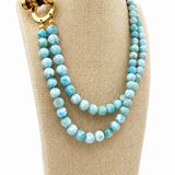 A double-strand beaded necklace with light blue marbled beads and a gold-toned clasp, displayed on a beige fabric jewelry bust against a white background.