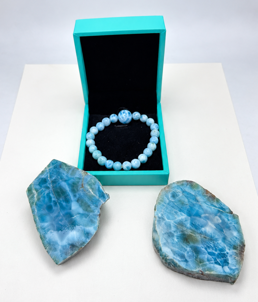 A light blue beaded bracelet with a central charm, displayed inside an open turquoise jewelry box, with two blue marbled stone slabs on a white surface against a light gray background.