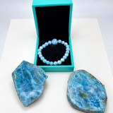 A light blue beaded bracelet with a central charm, displayed inside an open turquoise jewelry box, with two blue marbled stone slabs on a white surface against a light gray background.