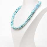 A Larimar bead
 necklace made of small irregular shell-like pieces in light blue and aqua tones, displayed on a white jewelry bust against a clean white background.
