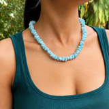 A woman wearing a chunky light blue beaded necklace and a dark teal ribbed tank top, photographed outdoors against a blurred green foliage background.