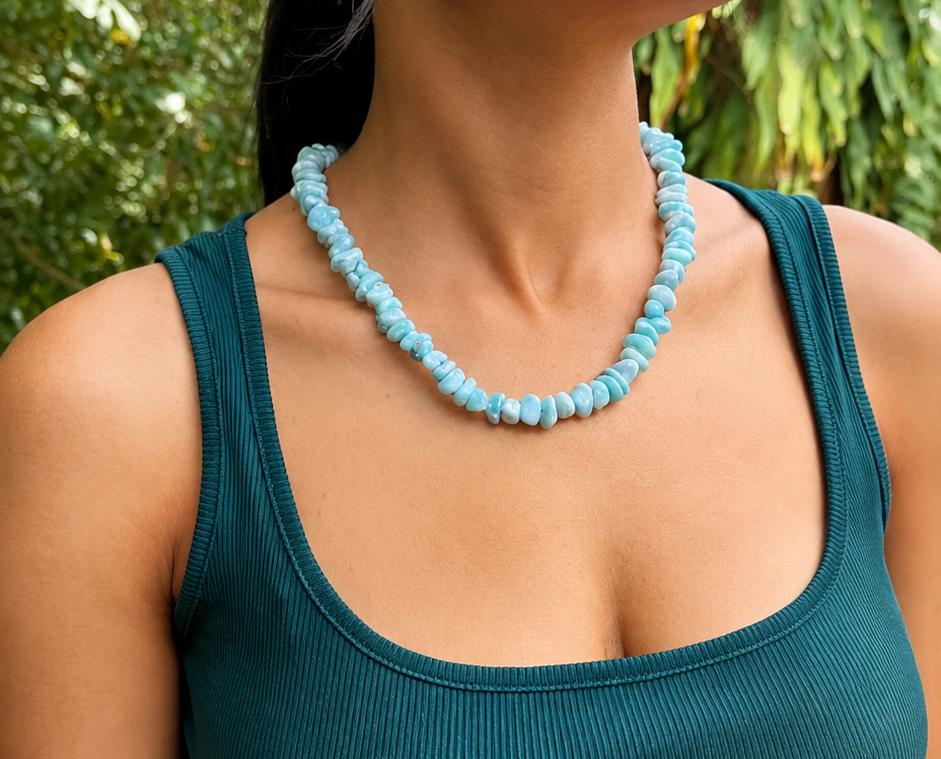 A woman wearing a chunky light blue beaded necklace and a dark teal ribbed tank top, photographed outdoors against a blurred green foliage background.
