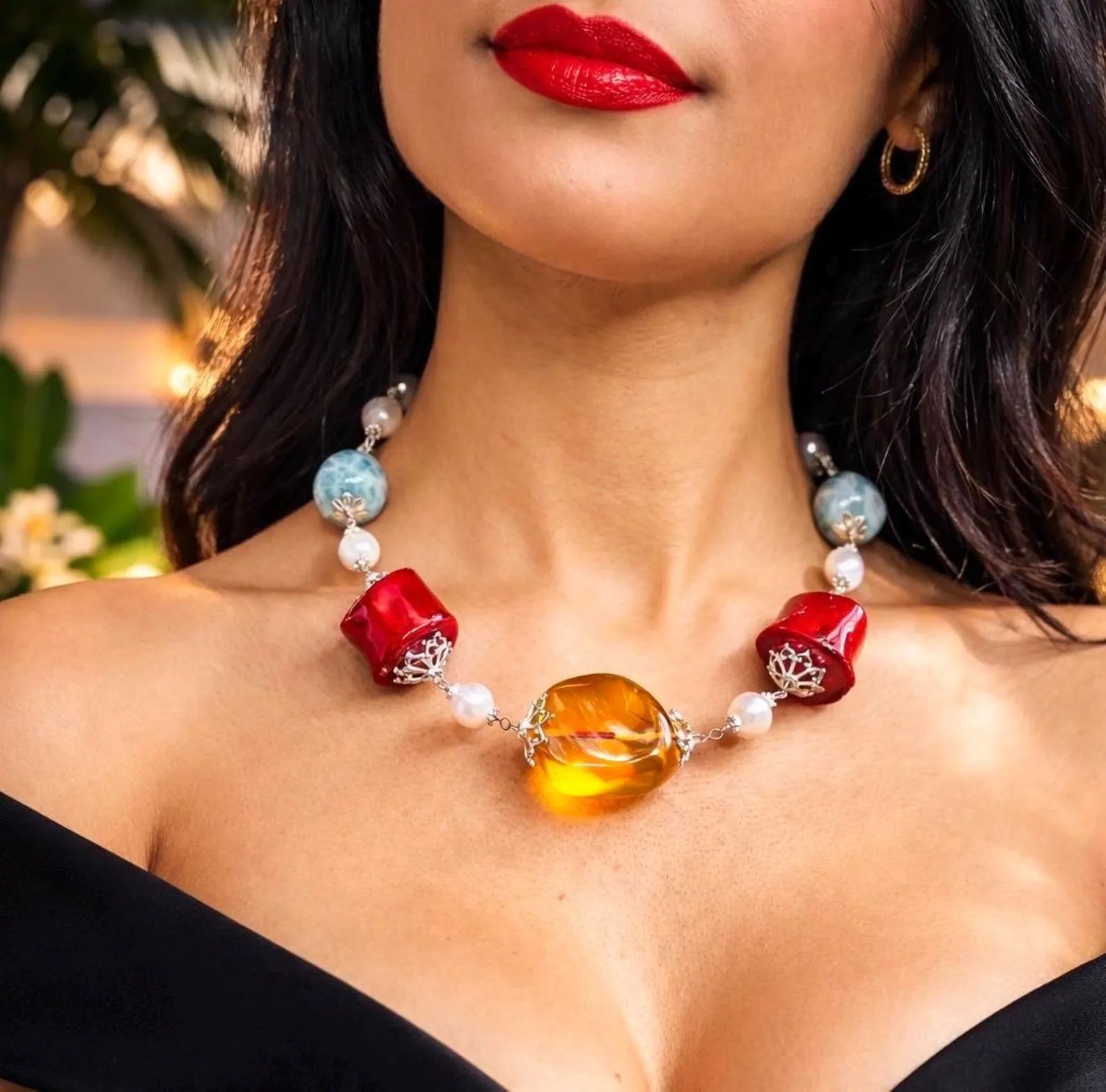 A close-up lifestyle shot of a woman wearing a colorful glass bead necklace. The necklace features a large, polished amber-yellow oval centerpiece, two red cylindrical beads with silver filigree details, white pearl-like beads, and light blue round beads.