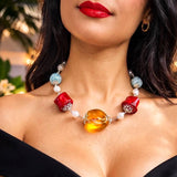 A close-up lifestyle shot of a woman wearing a colorful glass bead necklace. The necklace features a large, polished amber-yellow oval centerpiece, two red cylindrical beads with silver filigree details, white pearl-like beads, and light blue round beads.