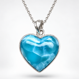 A Larimar Stone heart-shaped pendant necklace featuring a vibrant blue marbled gemstone in a polished silver bezel, suspended from a matching silver chain against a light gray background.