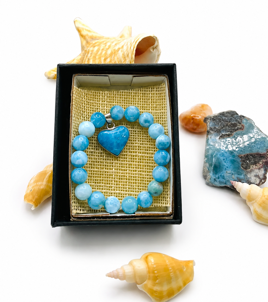 A beaded bracelet with light blue and medium blue round beads and a heart-shaped blue charm, presented in an open black gift box with a gold mesh insert. The box is styled on a white background with seashells and blue stones around it.