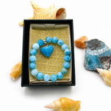 A beaded bracelet with light blue and medium blue round beads and a heart-shaped blue charm, presented in an open black gift box with a gold mesh insert. The box is styled on a white background with seashells and blue stones around it.