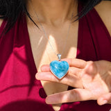 A silver heart-shaped pendant necklace with a vibrant blue marbled stone, held by a person's hand against a dark red halter top.