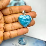 A heart-shaped larimar pendant in a silver bezel setting, held in a person's hand. A 'Mauna Vibes Certificate of Authenticity' card is visible in the blurred background.