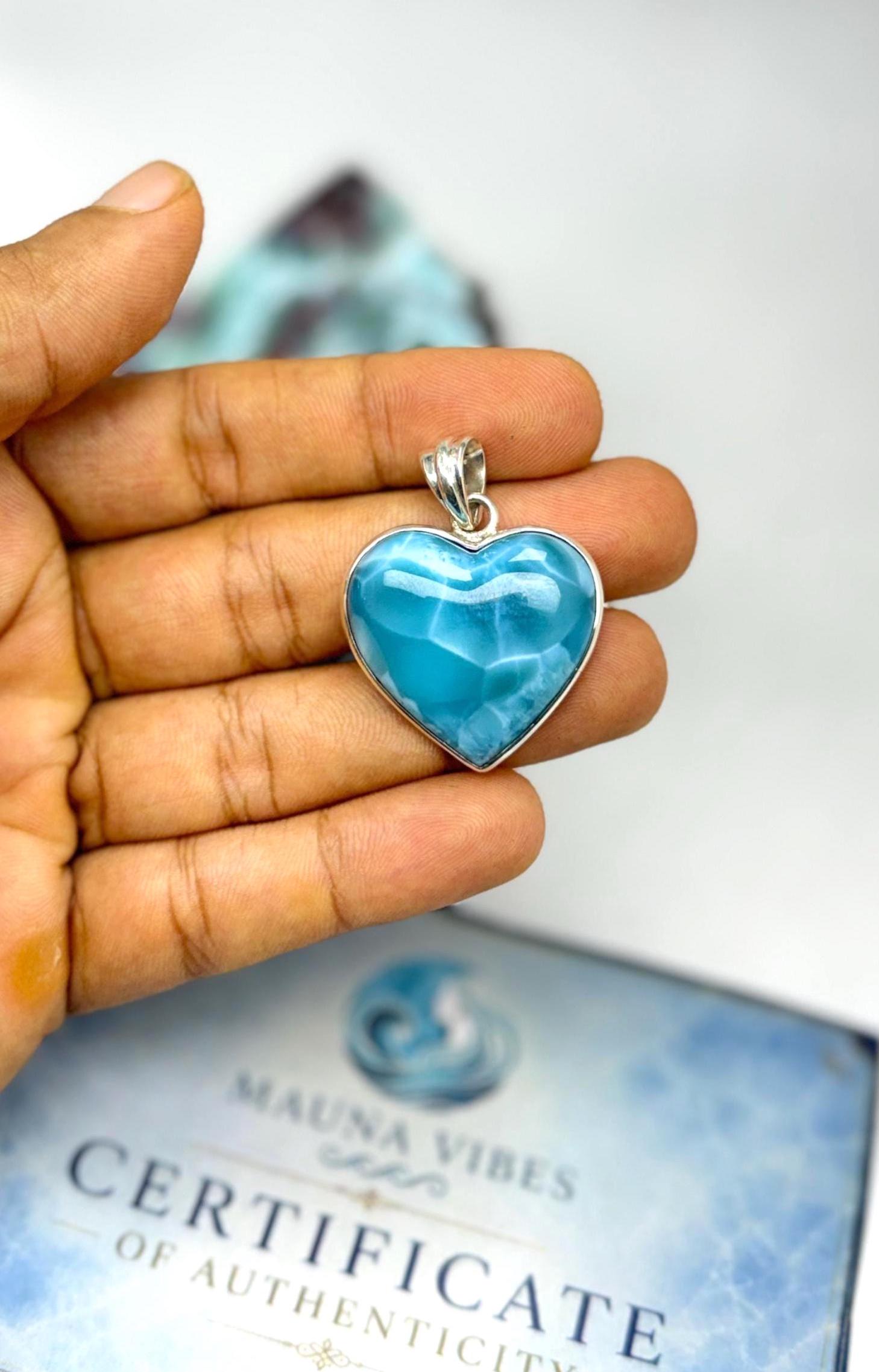 A heart-shaped larimar pendant in a silver bezel setting, held in a person's hand. A 'Mauna Vibes Certificate of Authenticity' card is visible in the blurred background.
