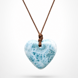 A Larimar heart-shaped pendant made of light blue and white marbled stone, suspended from a brown cord necklace against a clean white background.