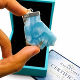 A hand holds a larimar stone pendant with a silver bail in front of an open turquoise box. A certificate card reading 'MAUNA VIBES CERTIFICATE AUTHENTICITY' is visible on a white background.