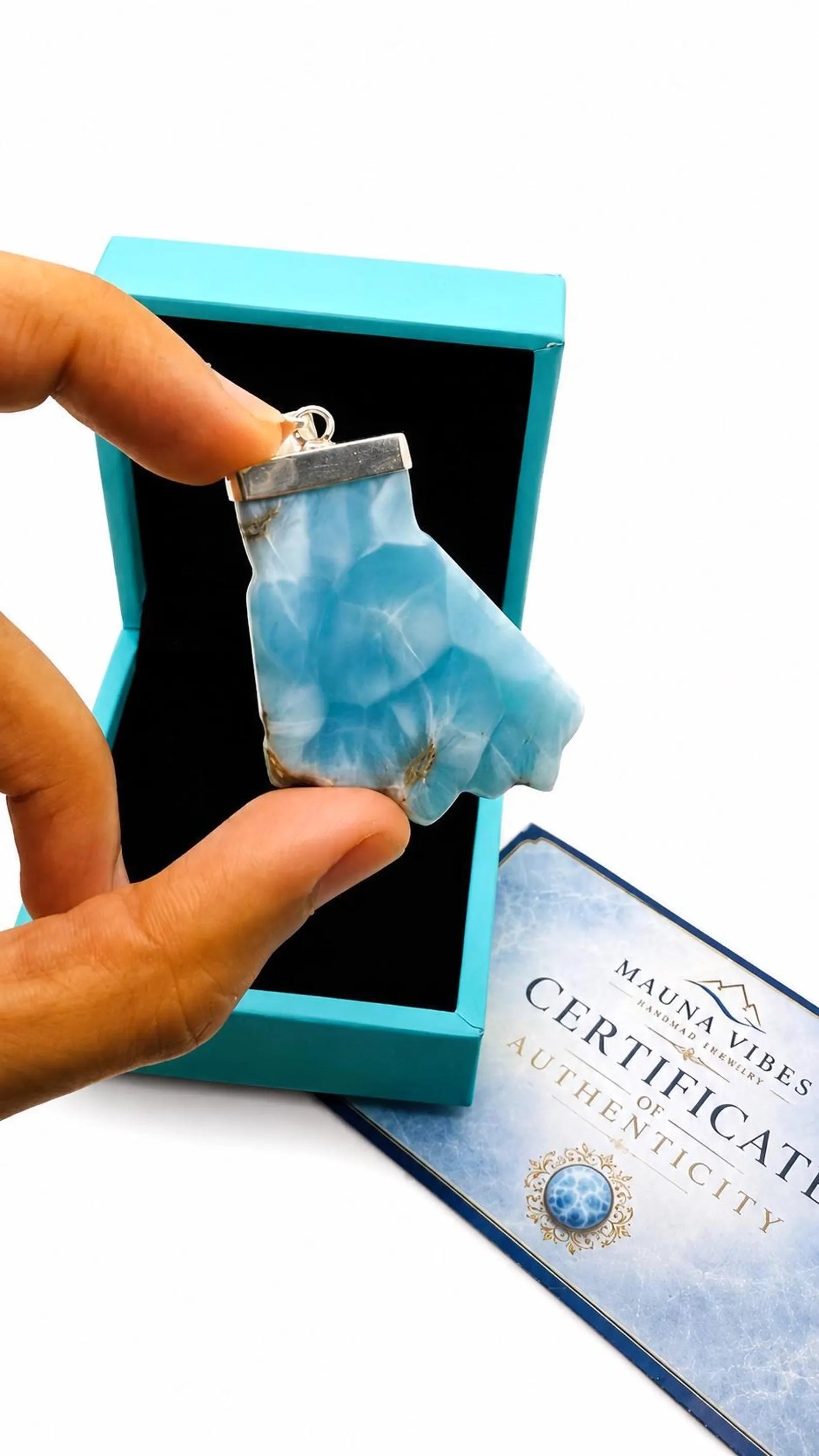 A hand holds a larimar stone pendant with a silver bail in front of an open turquoise box. A certificate card reading 'MAUNA VIBES CERTIFICATE AUTHENTICITY' is visible on a white background.