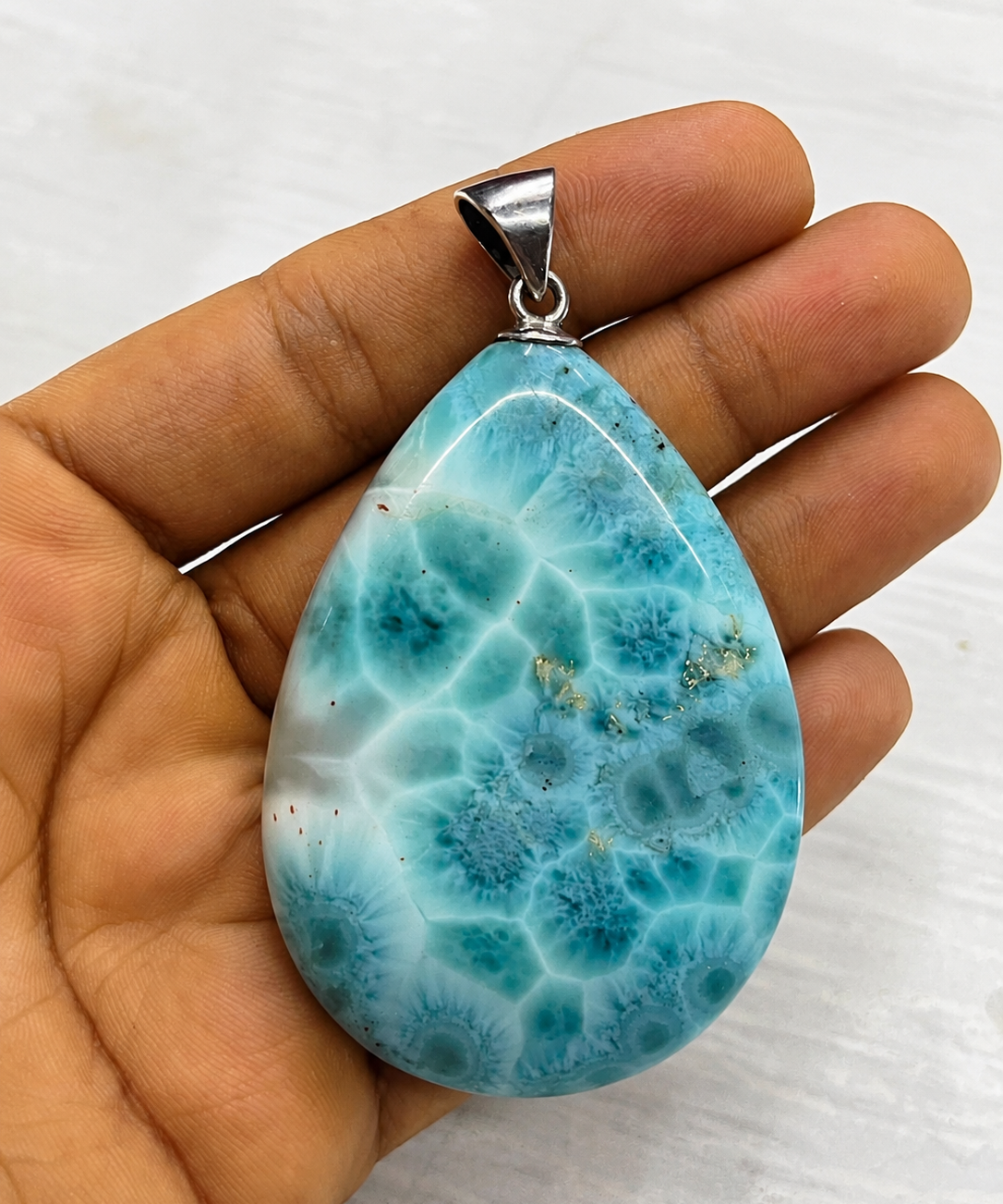 A  Larimar teardrop-shaped pendant made of polished turquoise-blue and white marbled stone, held in a person's hand against a light gray background.