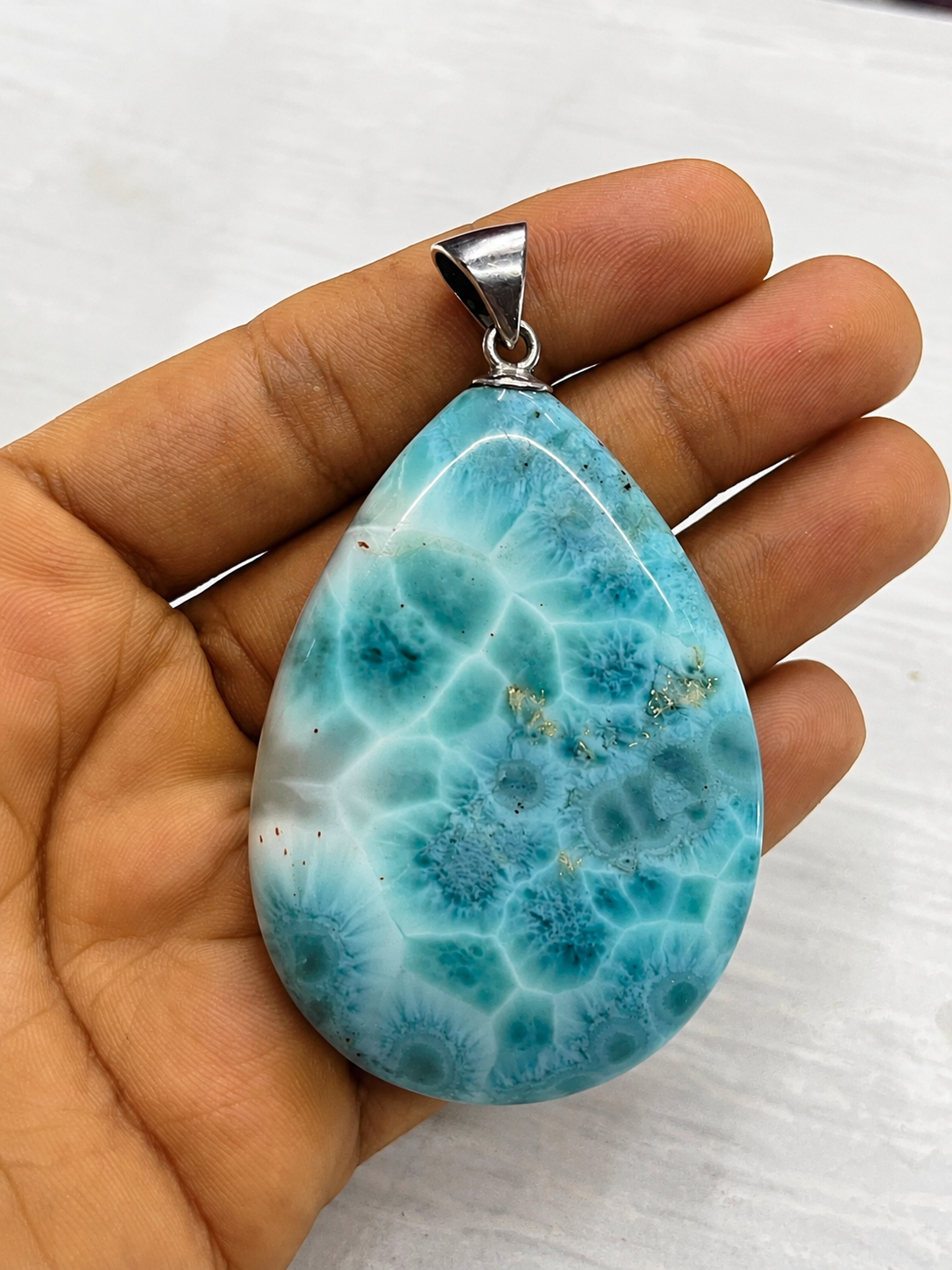 A  Larimar teardrop-shaped pendant made of polished turquoise-blue and white marbled stone, held in a person's hand against a light gray background.