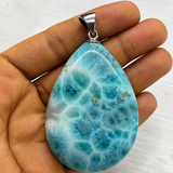 A  Larimar teardrop-shaped pendant made of polished turquoise-blue and white marbled stone, held in a person's hand against a light gray background.