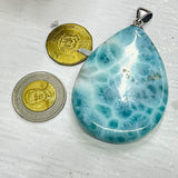 A teardrop-shaped labradorite pendant with a marbled blue and white pattern, shown next to two coins for scale on a textured white surface.