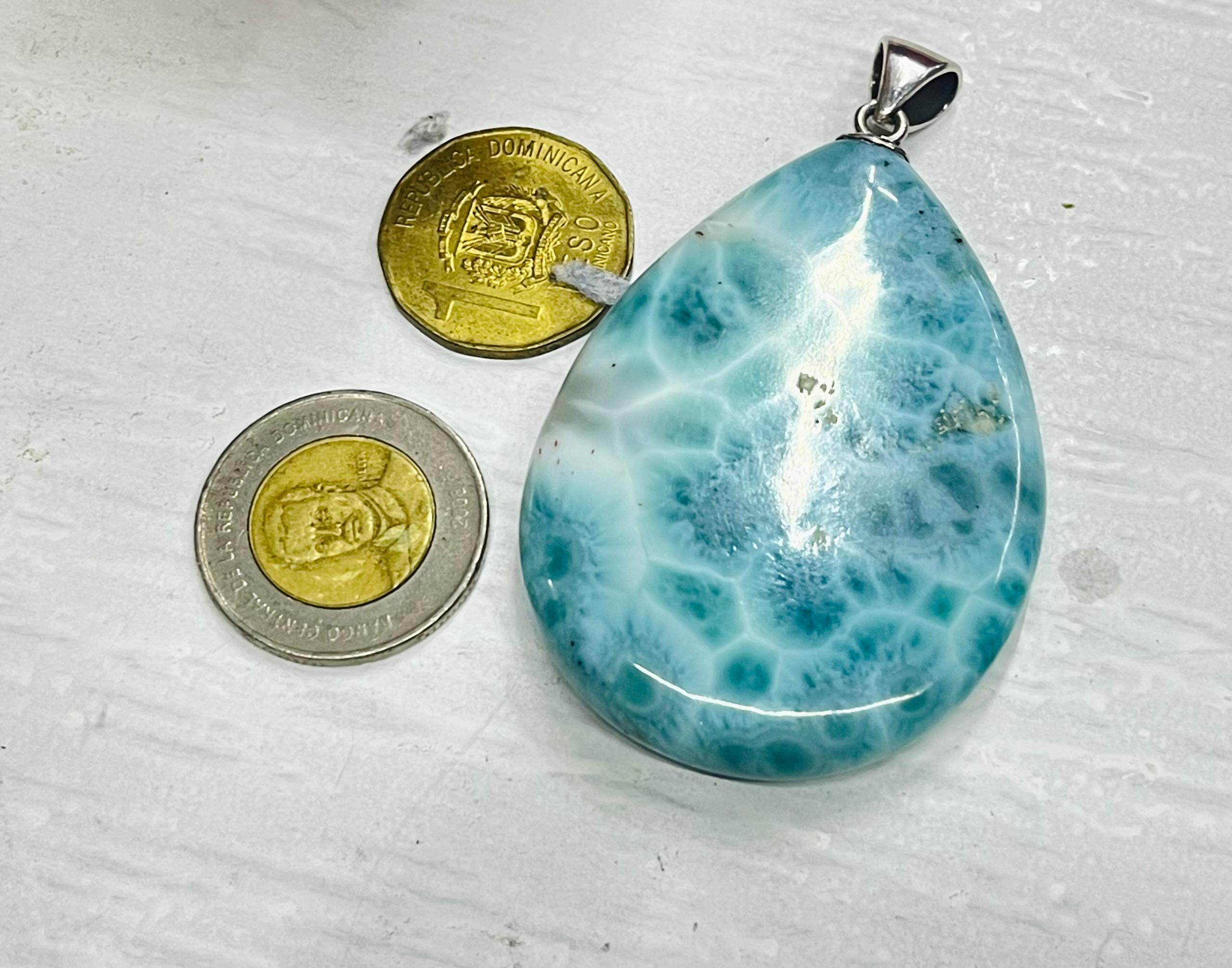 A teardrop-shaped labradorite pendant with a marbled blue and white pattern, shown next to two coins for scale on a textured white surface.