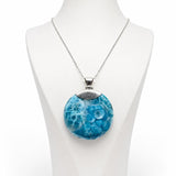 A silver chain larimar necklace with a round pendant featuring a textured blue and white pattern, displayed on a white mannequin bust against a white background.