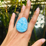 A close-up of a hand wearing a large teardrop-shaped light blue marbled stone ring with a silver-toned bezel, set against a blurred natural background of green foliage.