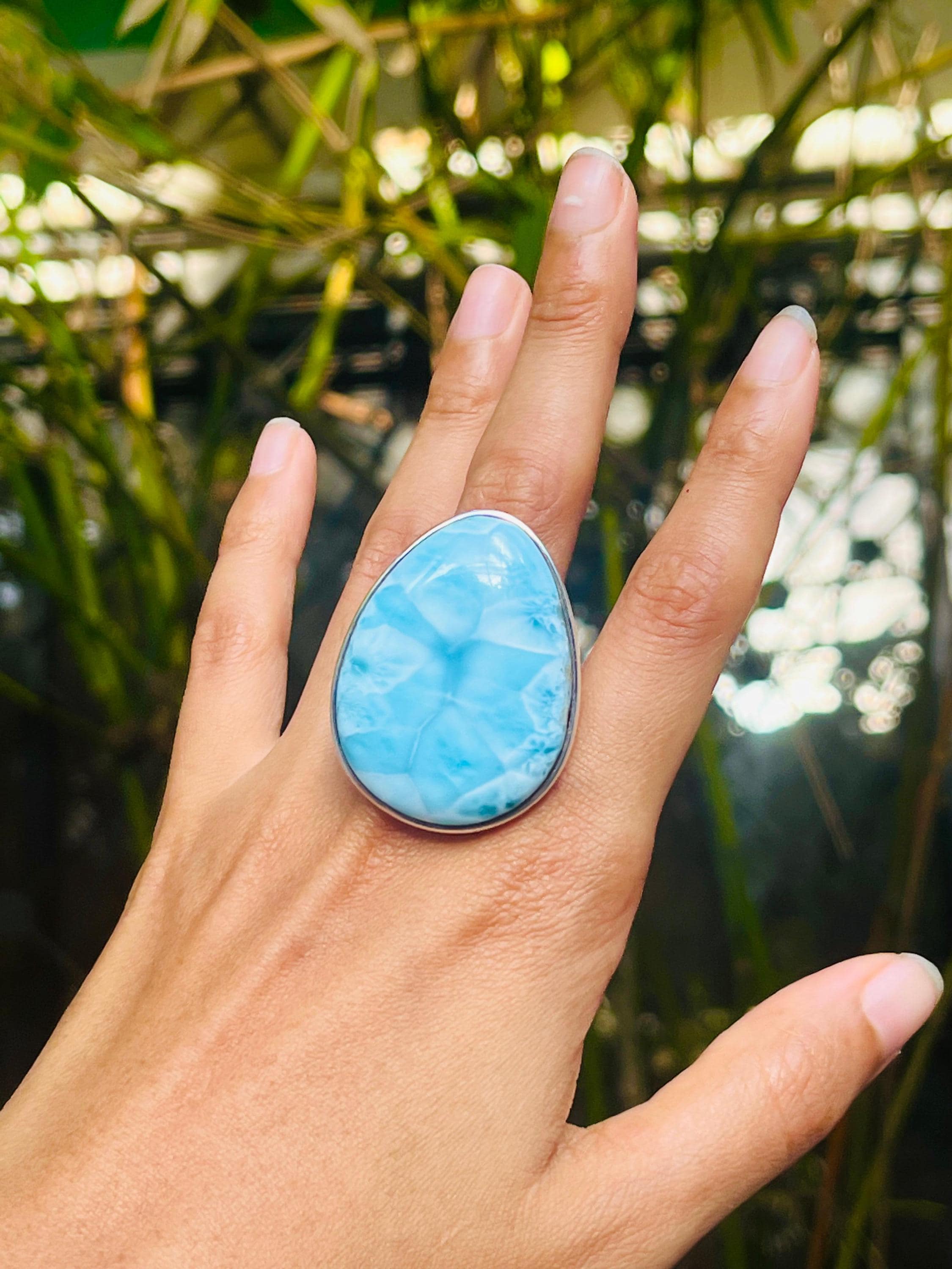 A close-up of a hand wearing a large teardrop-shaped light blue marbled stone ring with a silver-toned bezel, set against a blurred natural background of green foliage.