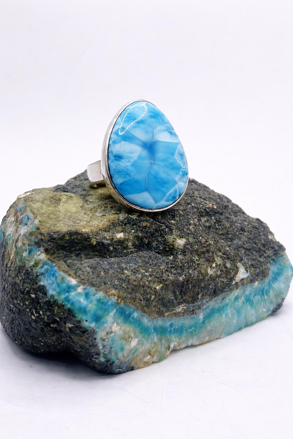 A silver LaRIMAR 
 ring with a large oval blue marbled gemstone, displayed on a rough turquoise and green stone against a white background.