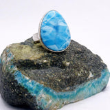 A silver LaRIMAR 
 ring with a large oval blue marbled gemstone, displayed on a rough turquoise and green stone against a white background.
