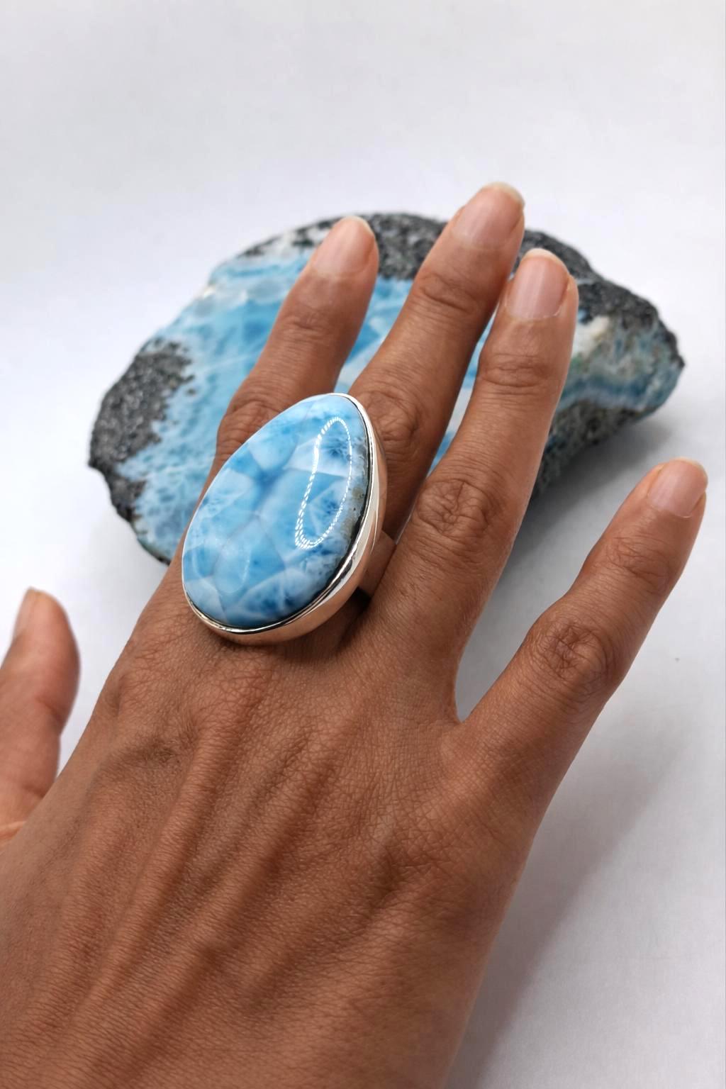 A hand wearing a large ring with a blue and white marbled stone, set in a silver-toned bezel. A blue and white geode is displayed in the background on a white surface.