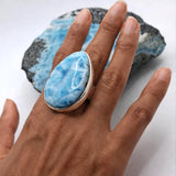 A hand wearing a large ring with a blue and white marbled stone, set in a silver-toned bezel. A blue and white geode is displayed in the background on a white surface.