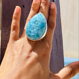 A close-up of a hand wearing a large teardrop-shaped turquoise ring with a silver-toned setting, posed against a dark wooden post with a beach background.