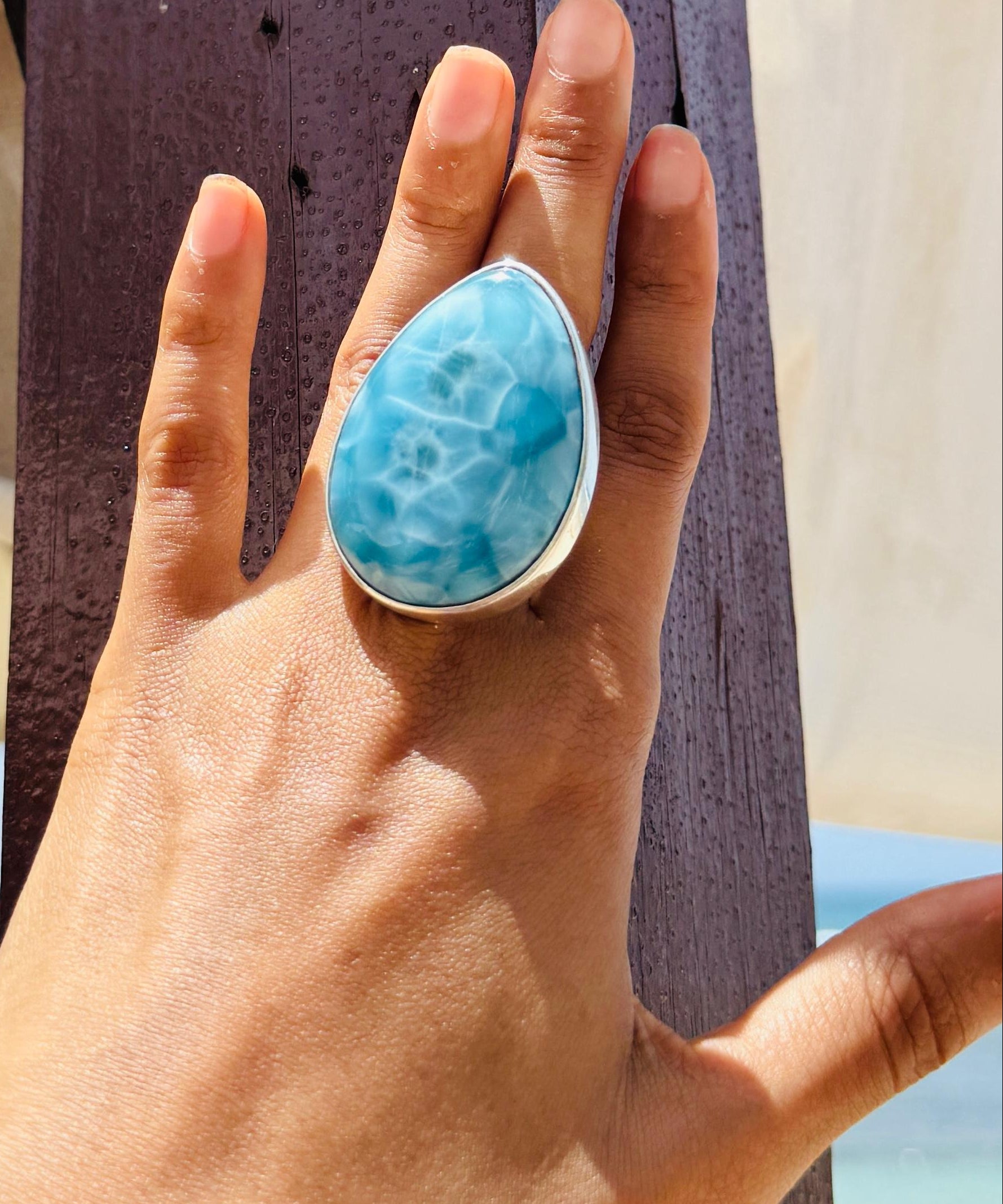 A close-up of a hand wearing a large teardrop-shaped turquoise ring with a silver-toned setting, posed against a dark wooden post with a beach background.