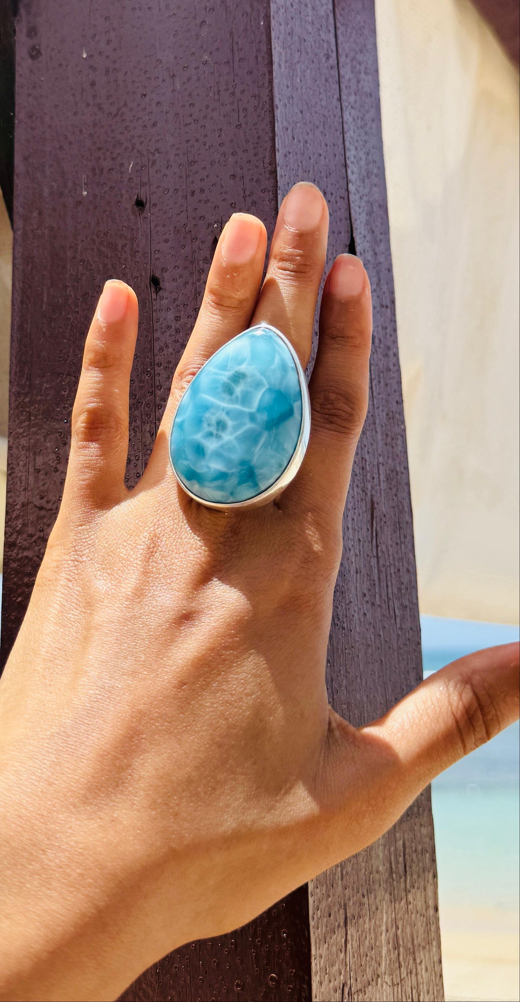 A close-up of a hand wearing a large teardrop-shaped turquoise ring with a silver-toned setting, posed against a dark wooden post with a beach background.