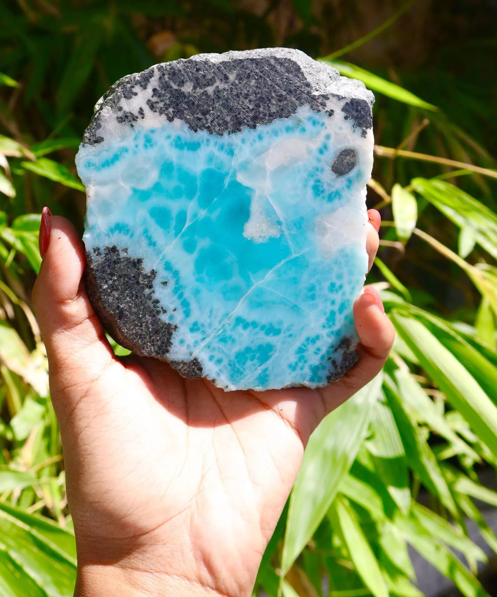 A hand holding a raw, polished baroque larimar gemstone featuring vibrant turquoise blue calcite crystals on a dark gray and white matrix, set against a blurred background of green foliage.
