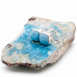 A pair of silver stud earrings with smooth, polished light blue gemstones, displayed on a natural blue and white mineral rock slice against a white background.