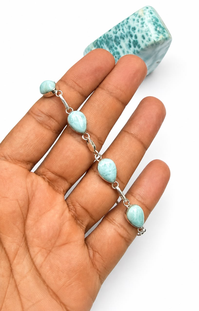 A silver chain bracelet with four light blue marbled teardrop beads and a matching bead at the end, displayed on a person's hand with a matching blue stone prop against a white background.