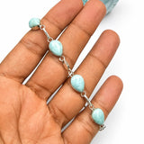 A silver chain bracelet with four light blue marbled teardrop beads and a matching bead at the end, displayed on a person's hand with a matching blue stone prop against a white background.