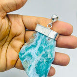A hand holding a raw, polished turquoise and white marbled gemstone pendant with a silver bezel setting, shown against a plain light gray background.