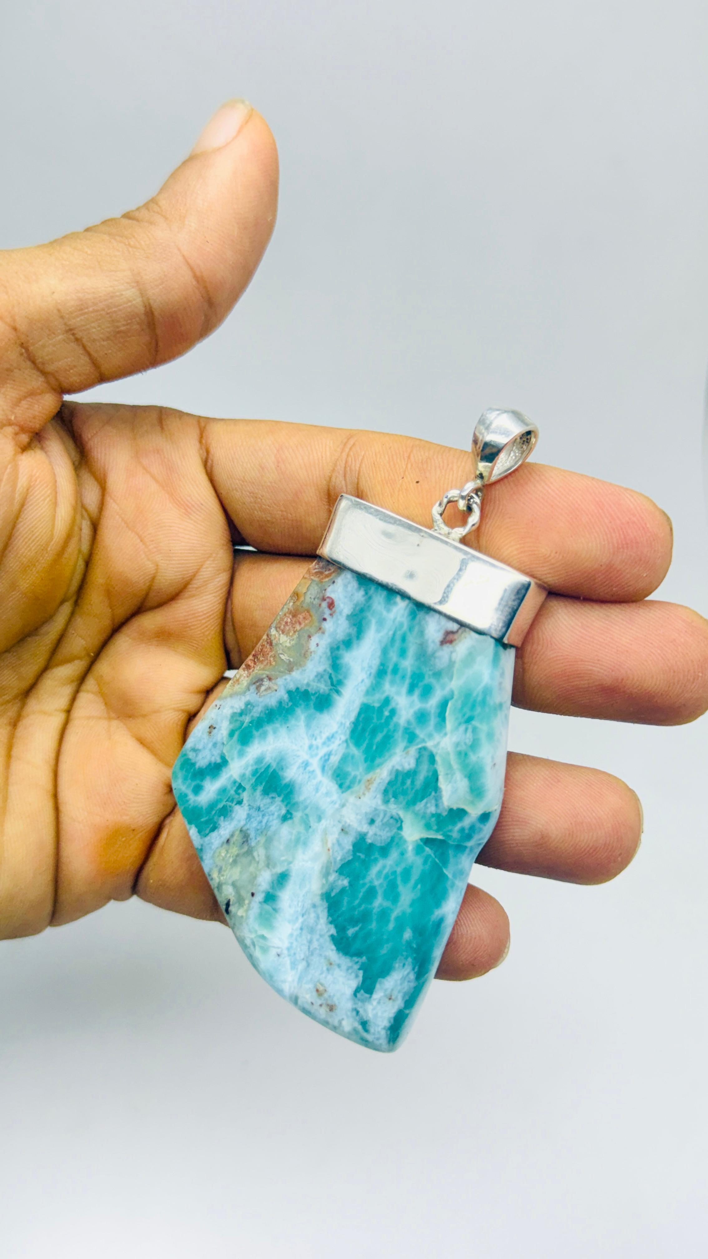A hand holding a raw, polished turquoise and white marbled gemstone pendant with a silver bezel setting, shown against a plain light gray background.