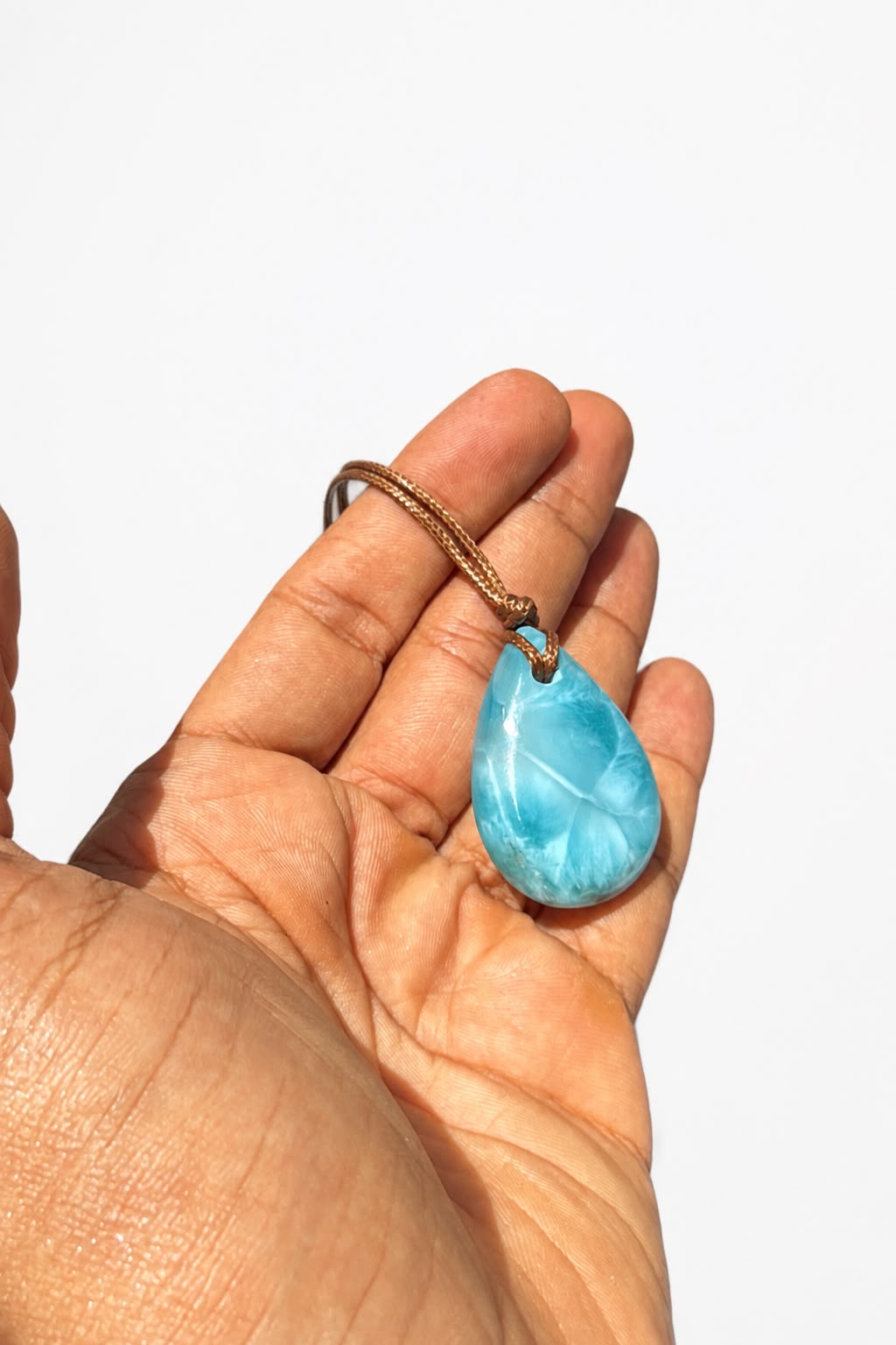 A hand holding a teardrop-shaped larimar pendant necklace with a brown cord, set against a plain white background.