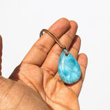 A hand holding a teardrop-shaped larimar pendant necklace with a brown cord, set against a plain white background.