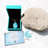 A larimar teardrop pendant necklace in a turquoise box, displayed with a coral shell, an authenticity card, and a certificate on a white surface with water-ripple shadows.