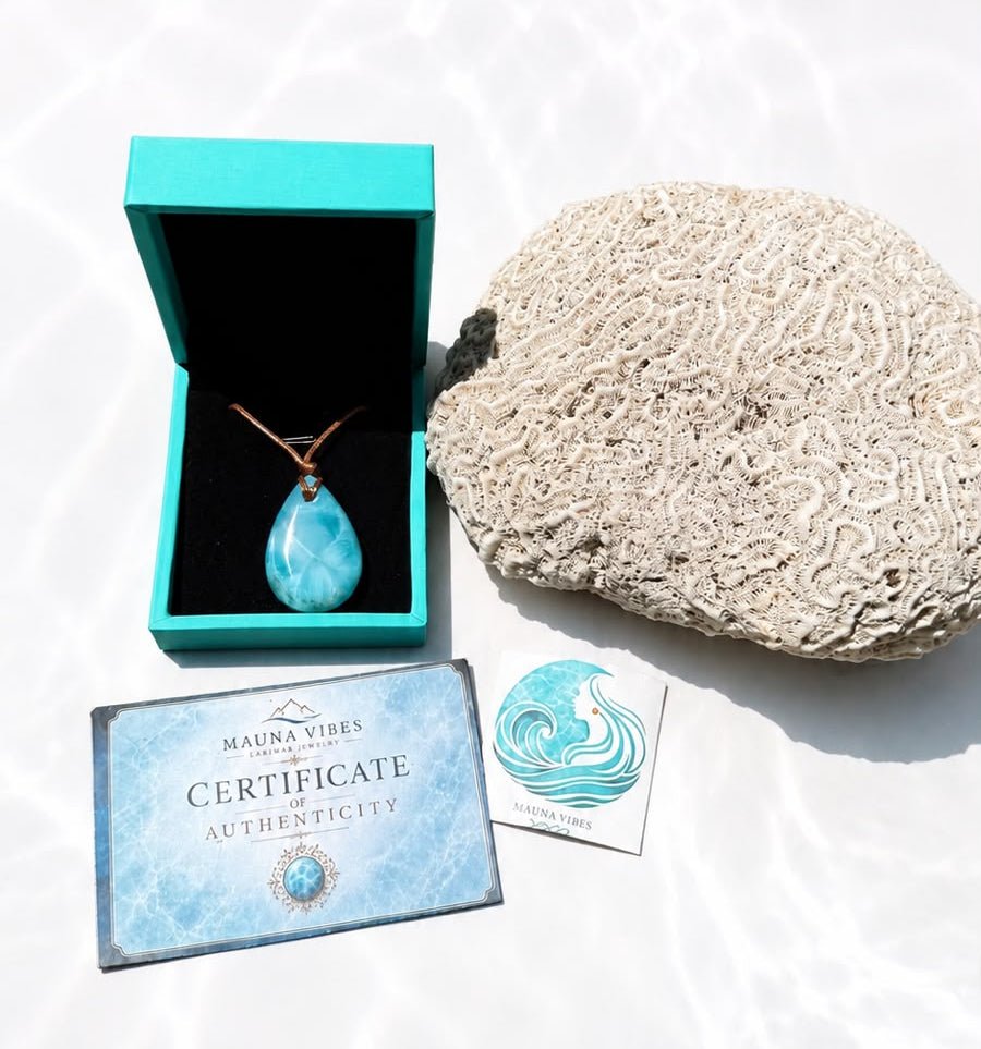 A larimar teardrop pendant necklace in a turquoise box, displayed with a coral shell, an authenticity card, and a certificate on a white surface with water-ripple shadows.