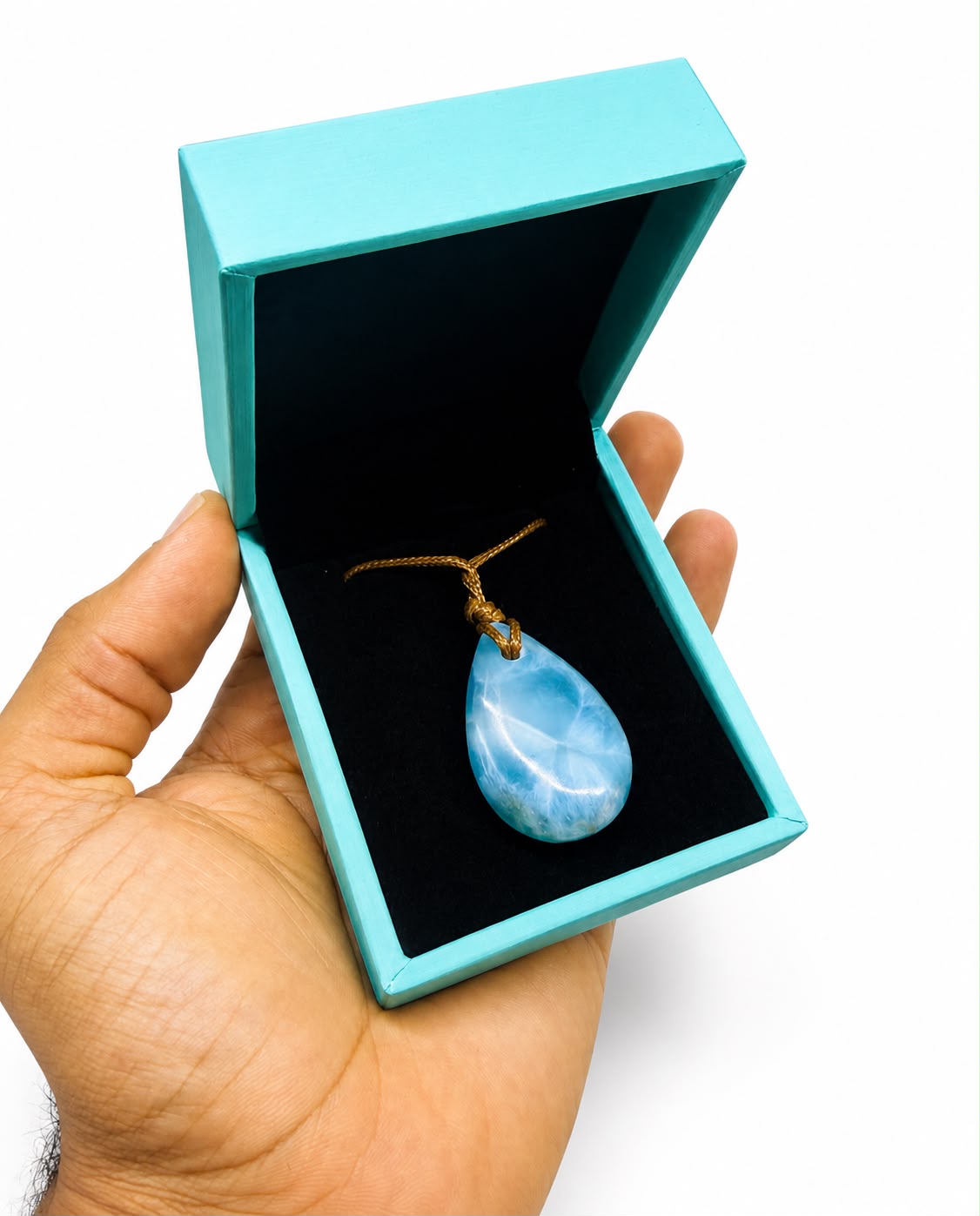 A hand holding an open turquoise jewelry box with a black velvet interior, displaying a teardrop-shaped larimar light blue stone pendant on a gold cord.