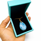A hand holding an open turquoise jewelry box with a black velvet interior, displaying a teardrop-shaped larimar light blue stone pendant on a gold cord.