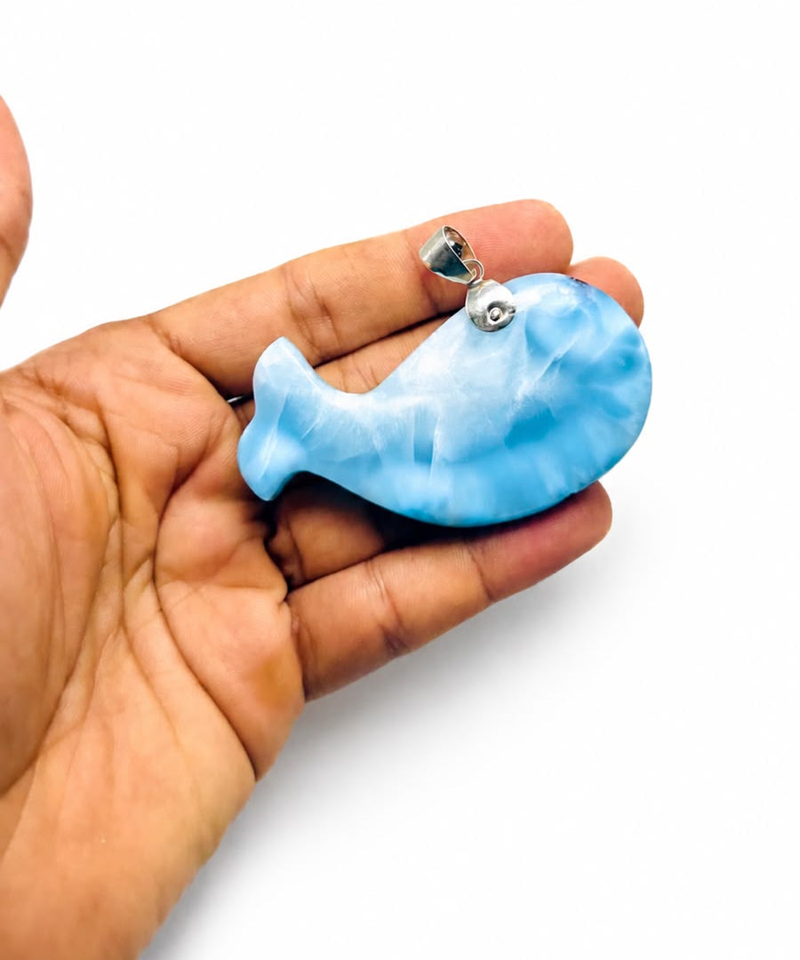 A hand holding a Larimar stone blue whale-shaped pendant with a silver bail, shown against a plain white background.