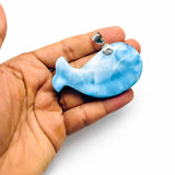 A hand holding a Larimar stone blue whale-shaped pendant with a silver bail, shown against a plain white background.