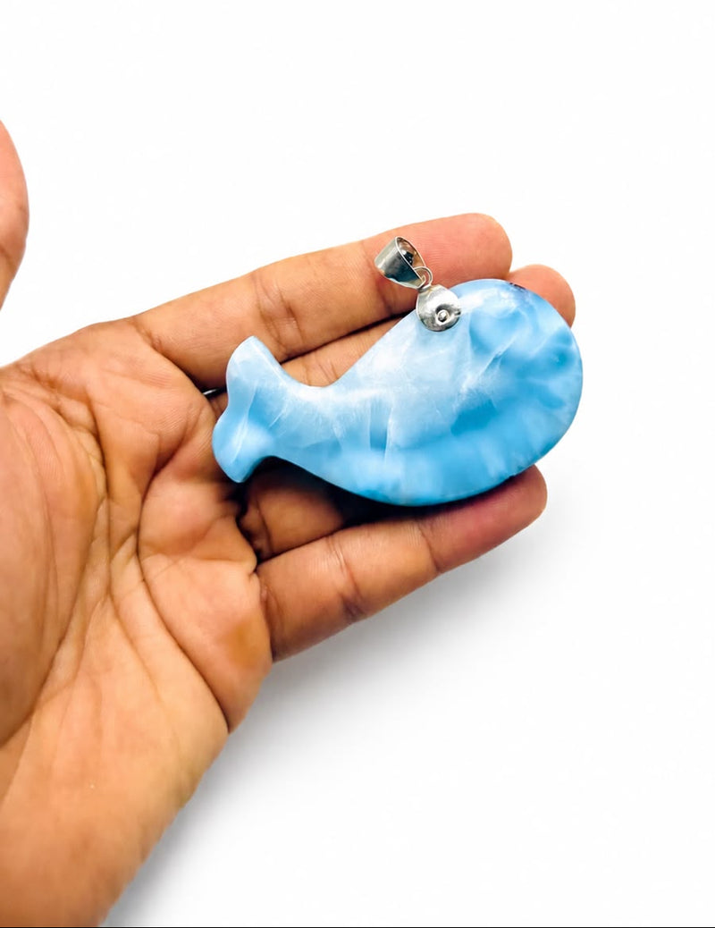 A hand holding a Larimar stone blue whale-shaped pendant with a silver bail, shown against a plain white background.