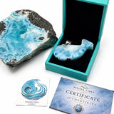 A larimar fish-shaped pendant displayed in an open turquoise box, alongside a large blue-and-black marbled stone, a small branded card, and a certificate card on a white background.